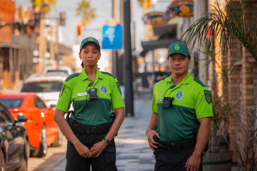Two Civicity Community Response Officers stand on a vibrant downtown street. One woman and one man wear bright green and dark green color blocked shirts with badges and body cameras with a green hat showing the Civicity logo